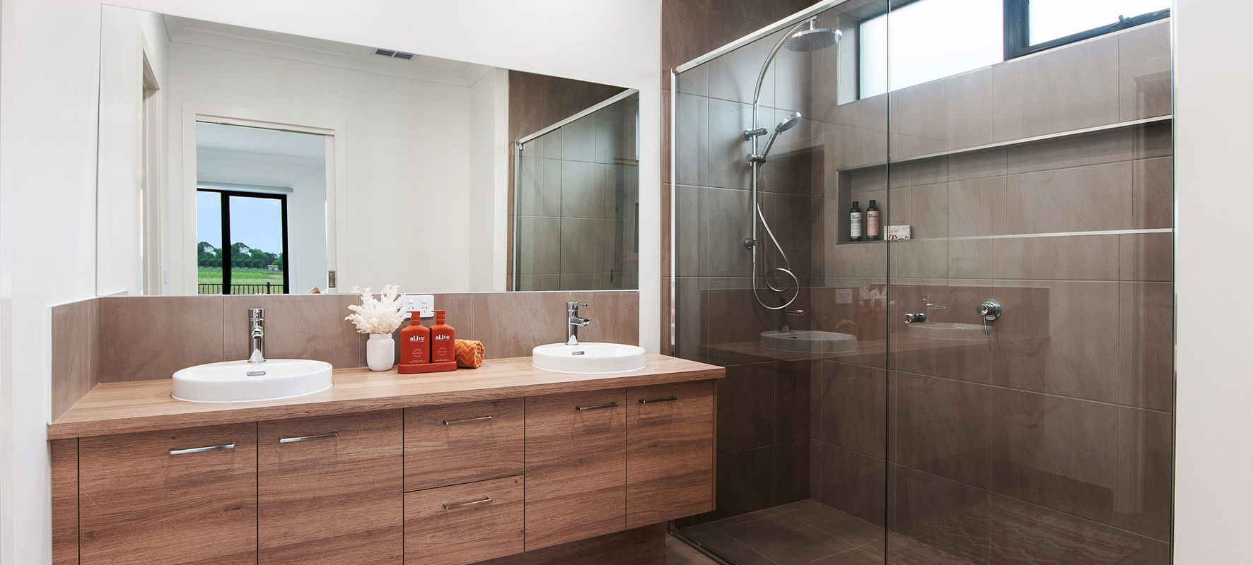 Photo of modern bathroom showing timber vanity with two white sinks, and clear glass shower screen with grey tiled shower