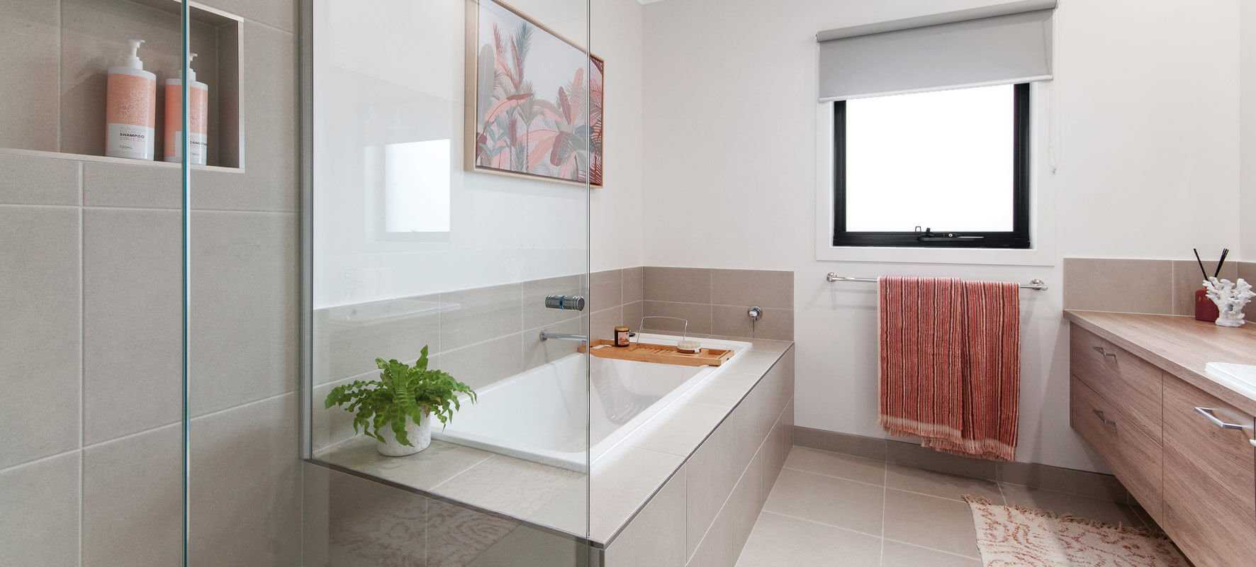 Photo of modern family bathroom with grey tiles, white bathub set in the tiles, timber vanity and clear glass shower screen. Pink floral painting on the wall.