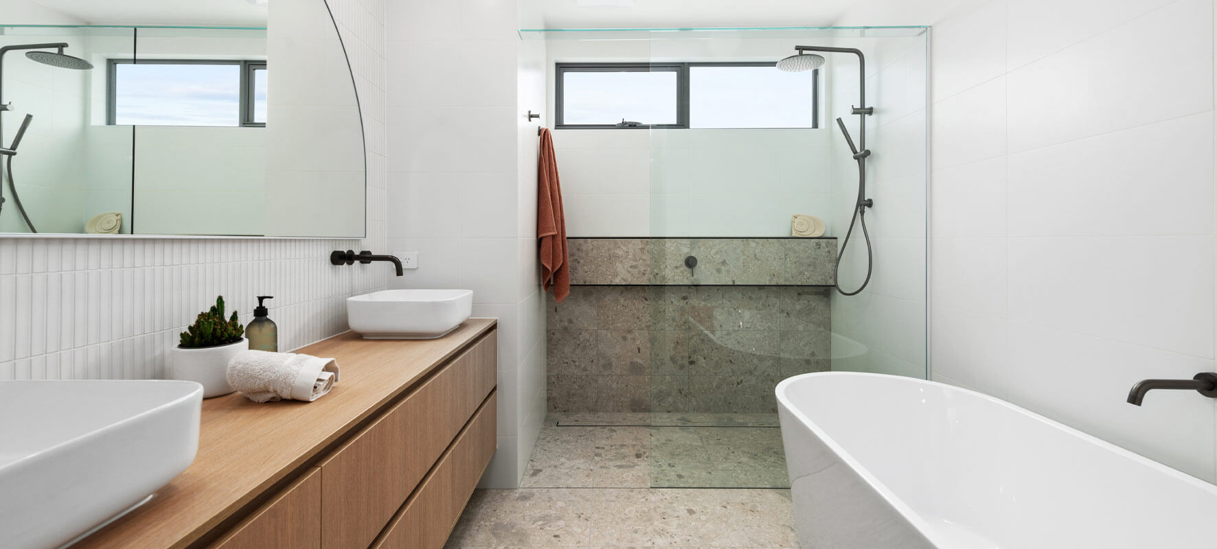 Photo of modern bathroom with white bathtub, timber vanity and white sink, grey tiled shower with clear glass shower screen