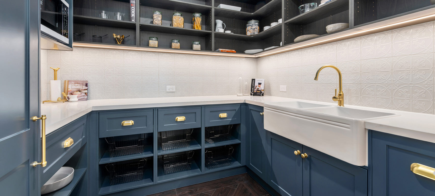Photo of butlers pantry in Ranch style home showing navy blue cabinets with white benchtops and feature white tiles. Gold tapware.