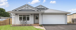 Photo of the facade of a home with dark grey colourbond tin roof, light grey weatherboards, white garage door and trims. The house has a pointed roof detail and verandah with a small garden at the front.