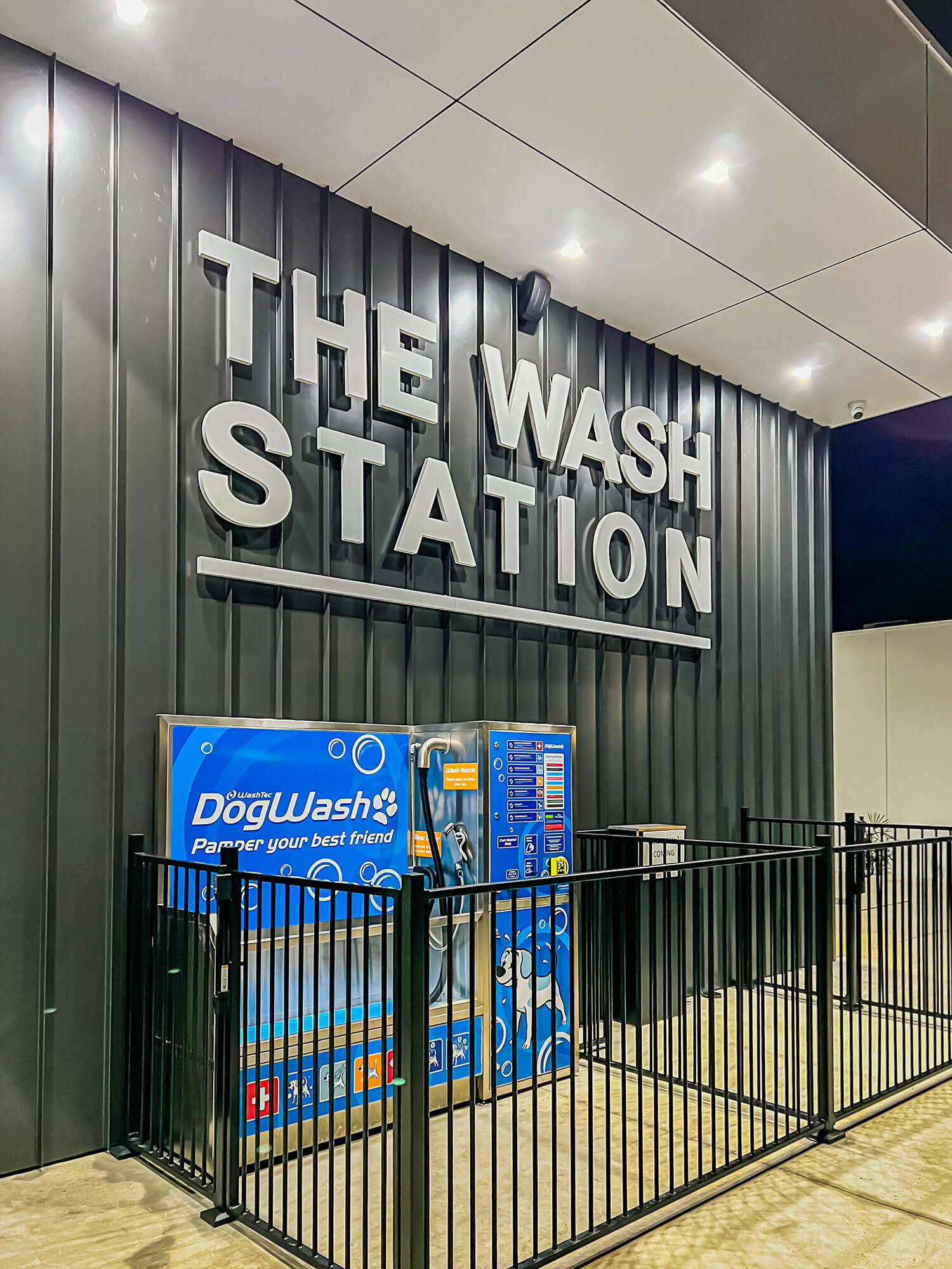 Photo of a large sign with white letters on a grey wall, the sign says 'The Wash Station'. Under the sign is a dog bath system with a black fence around it