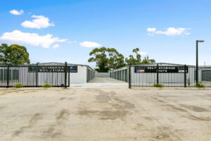 Photo of entry to a new storage shed facility, with black metal fencing and gates, and rows of grey storage shed with black roller doors