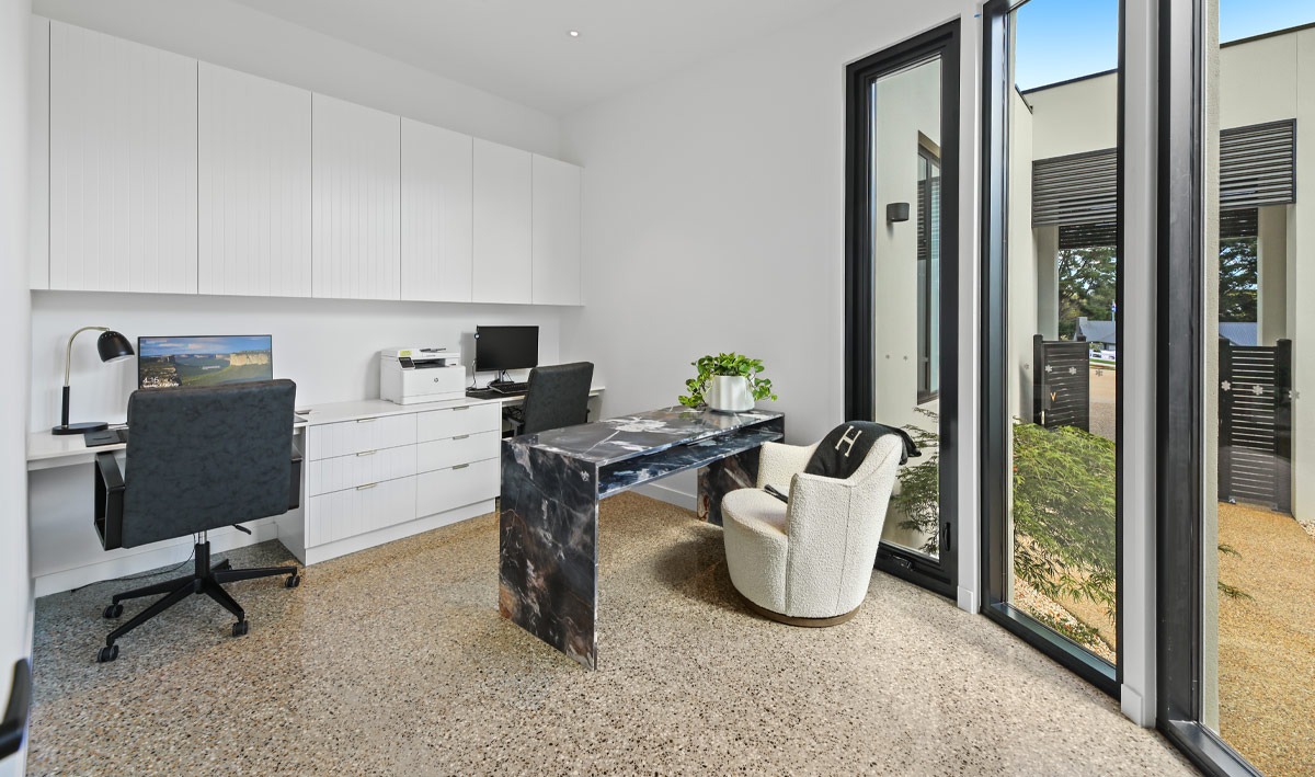 Working from home office space with polished concrete floor, white desk and cabinets along a wall, black leather chairs, and a large window to view a small garden