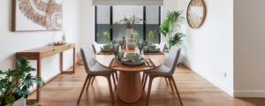 Photo of a wooden dining table with four chairs infront of a large sunny window. The table is set with placemats, cutlery, plates, bowls, cups and native flowers in the centre as a display. Chairs are fabric covered in cream. On the left is a woven artwork in a frame, a large clock is on the wall on the right. There are a few green plants in pots in the room, the floor is timber floorboards.