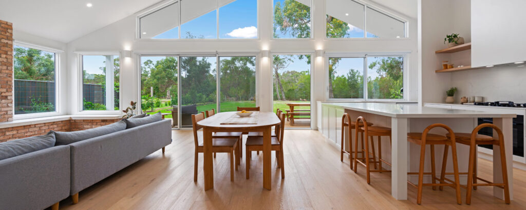 Inside a modern beach house looking across a dining table and out the large windows. The house has a high gabled ceiling and windows are full height.