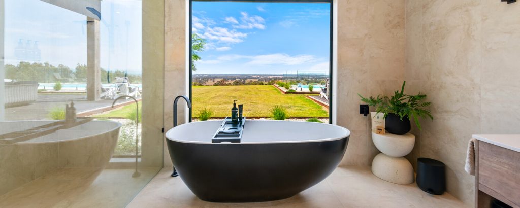 A peaceful image of a cream marble tile bathroom, with large black and white tub infront of a large picture window. View of blue sky and green farmland.