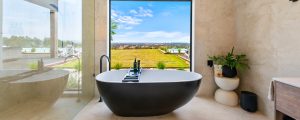 A peaceful image of a cream marble tile bathroom, with large black and white tub infront of a large picture window. View of blue sky and green farmland.
