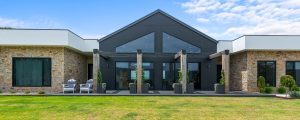 Facade of a large, modern farmhouse with stone walls on left and right that have a white flat roof, then in the centre of the home is a black feature gabled roofline with large windows.