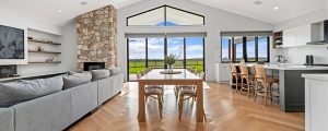 Open plan living area of a large, contemporary home with vaulted gable ceiling, shows grey lounge on the left, central timber dining setting seating 8, and grey and white kitchen on the right. Large windows look out onto green farmland.