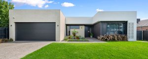 Facade of a contemporary home made of light grey brickwork and render, a flat roof, black garage door and windows, and feature garden of succulents and grasses.