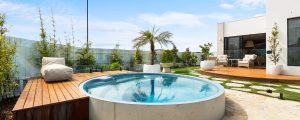 Photo of a round plungie pool set into the ground with tropical styl eplants around it and a white home with flat roof in the background.
