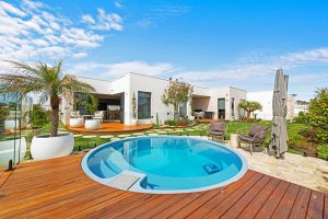 Blue sky above a contemporary new home with white bricks, flat roof, white render, and a large outdoor area with green grass and a round plunge pool in the foreground.