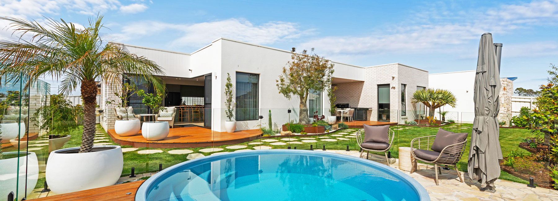 Blue sky above a contemporary new home with white bricks, flat roof, white render, and a large outdoor area with green grass and a round plunge pool in the foreground.